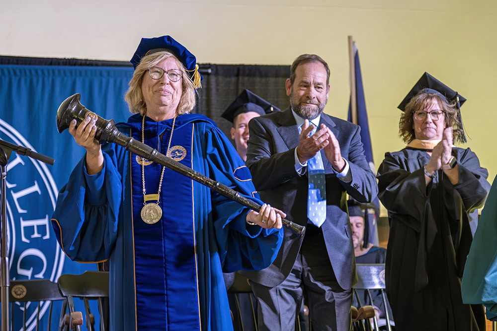 A woman in blue academic regalia holds a mace, flanked by a man and a woman in academic and formal attire who are clapping.