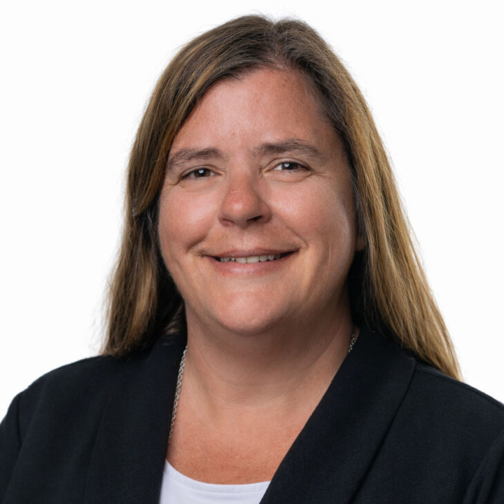 Headshot of a smiling woman with long brown hair, wearing a black blazer and a silver necklace against a white background.
